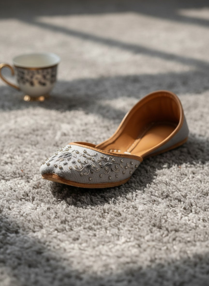 Ujaala Riwa gray jutti with intricate silver floral embroidery and bead embellishments on a light gray fabric.