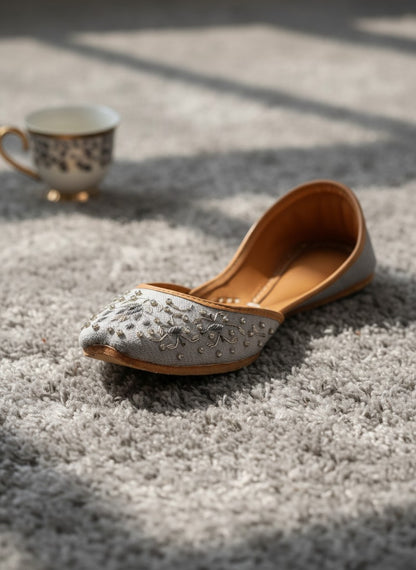 Ujaala Riwa gray jutti with intricate silver floral embroidery and bead embellishments on a light gray fabric.